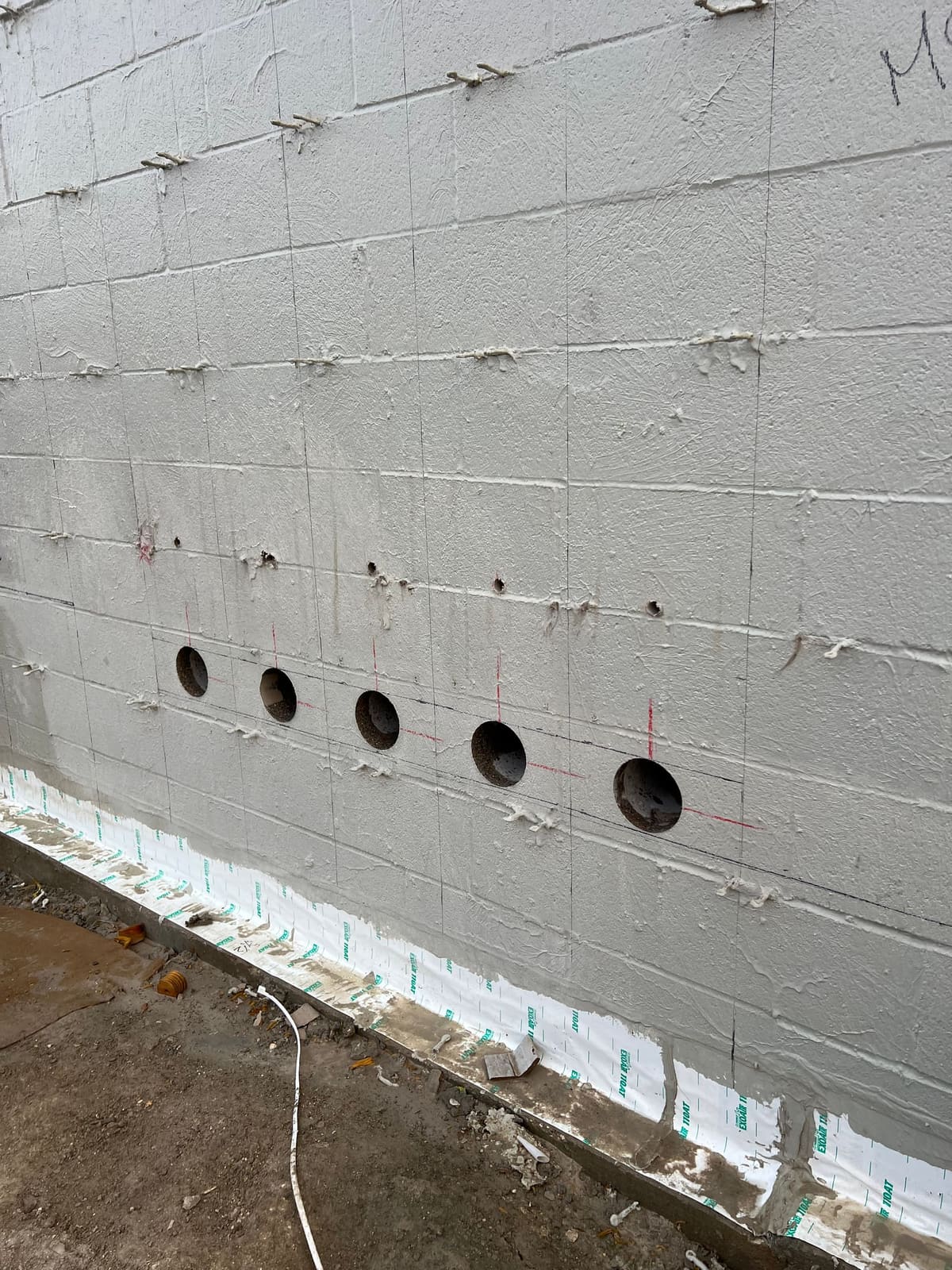 Row of angled core holes drilled through a painted CMU wall above a waterproofing membrane